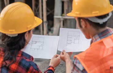 government contractors on a jobsite – DBL Center A man and a woman in hard hats on a job site, reading blueprints. Can only see the backs of their heads.