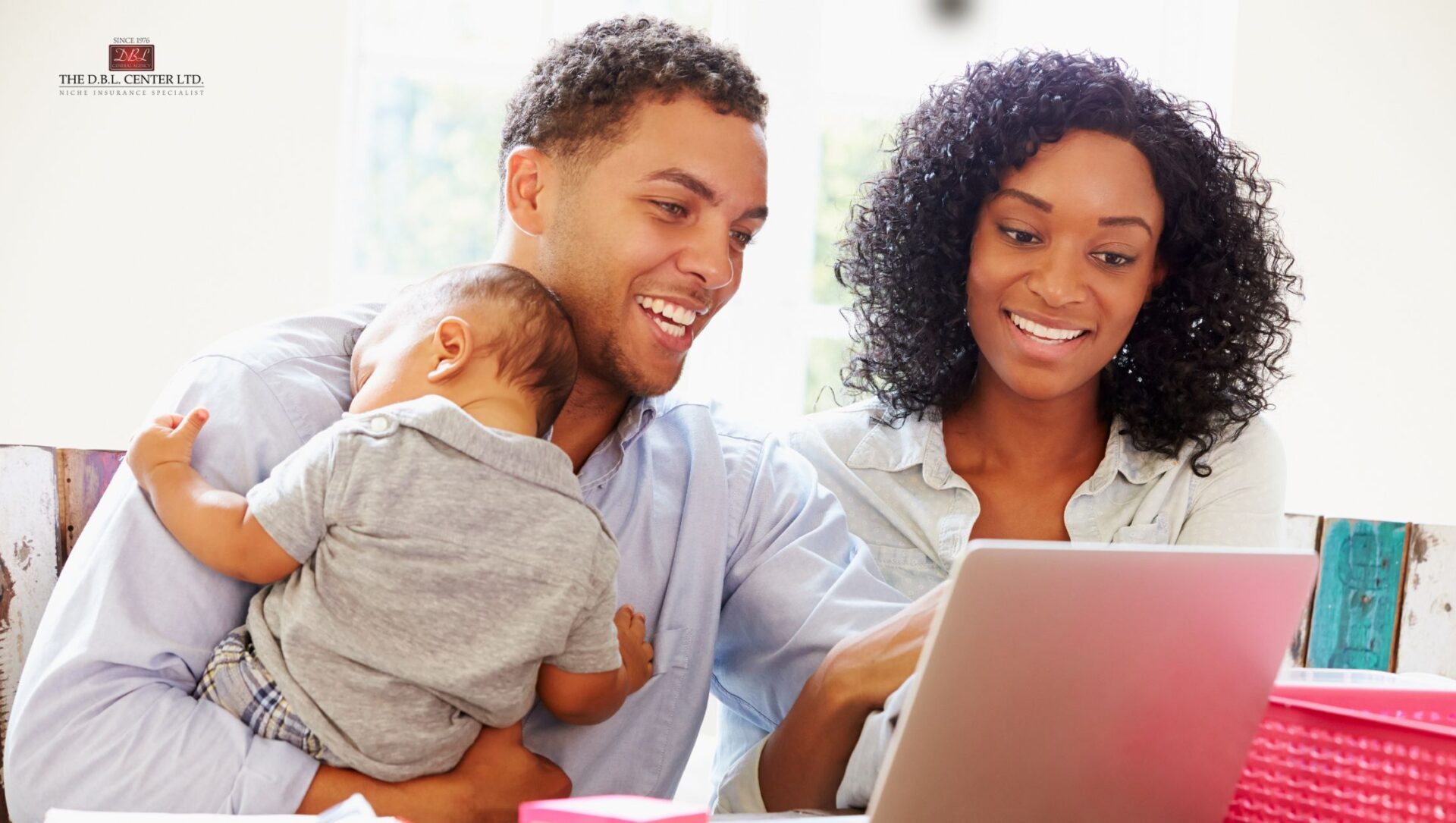 A couple holds an infant as they work together on a laptop computer.