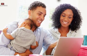 A couple holds an infant as they work together on a laptop computer.