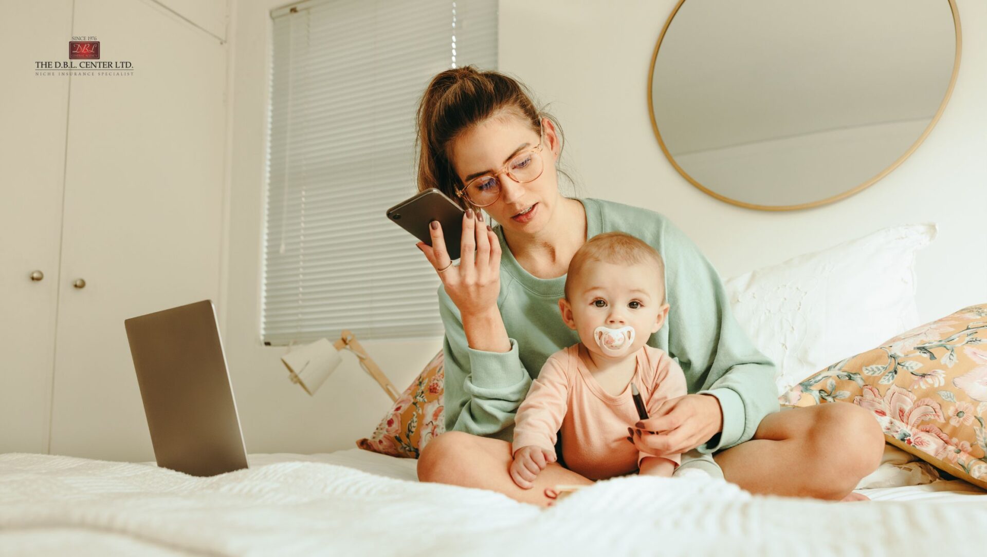 Mom with an infant on her lap, sitting on a bed, laptop open next to her. The mom is on her phone.