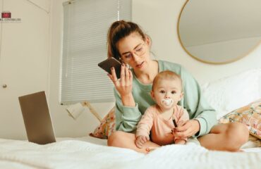 Mom with an infant on her lap, sitting on a bed, laptop open next to her. The mom is on her phone.